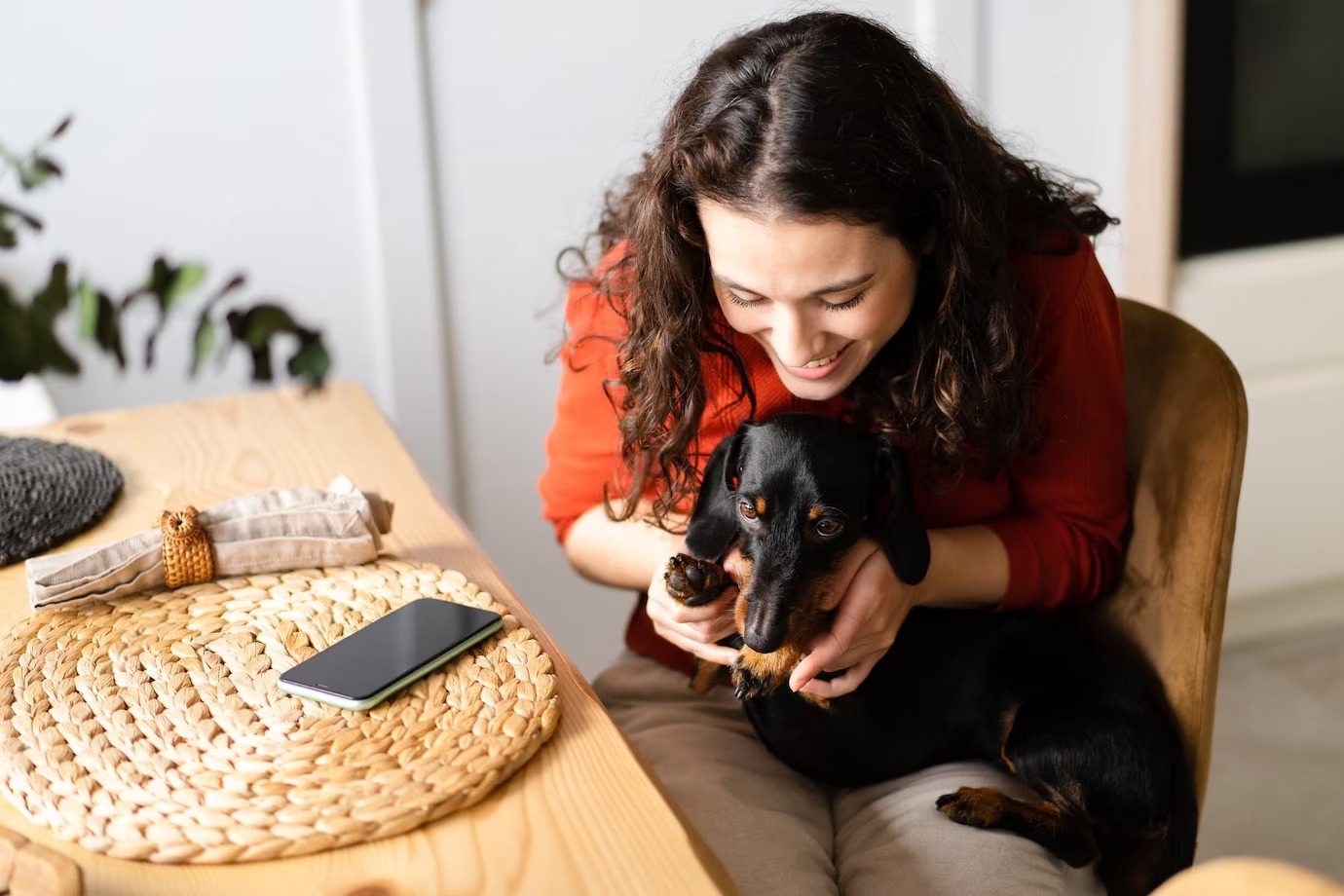A woman in a red sweater sits at a wooden table, holding a small black dachshund. A smartphone rests on the table beside them.