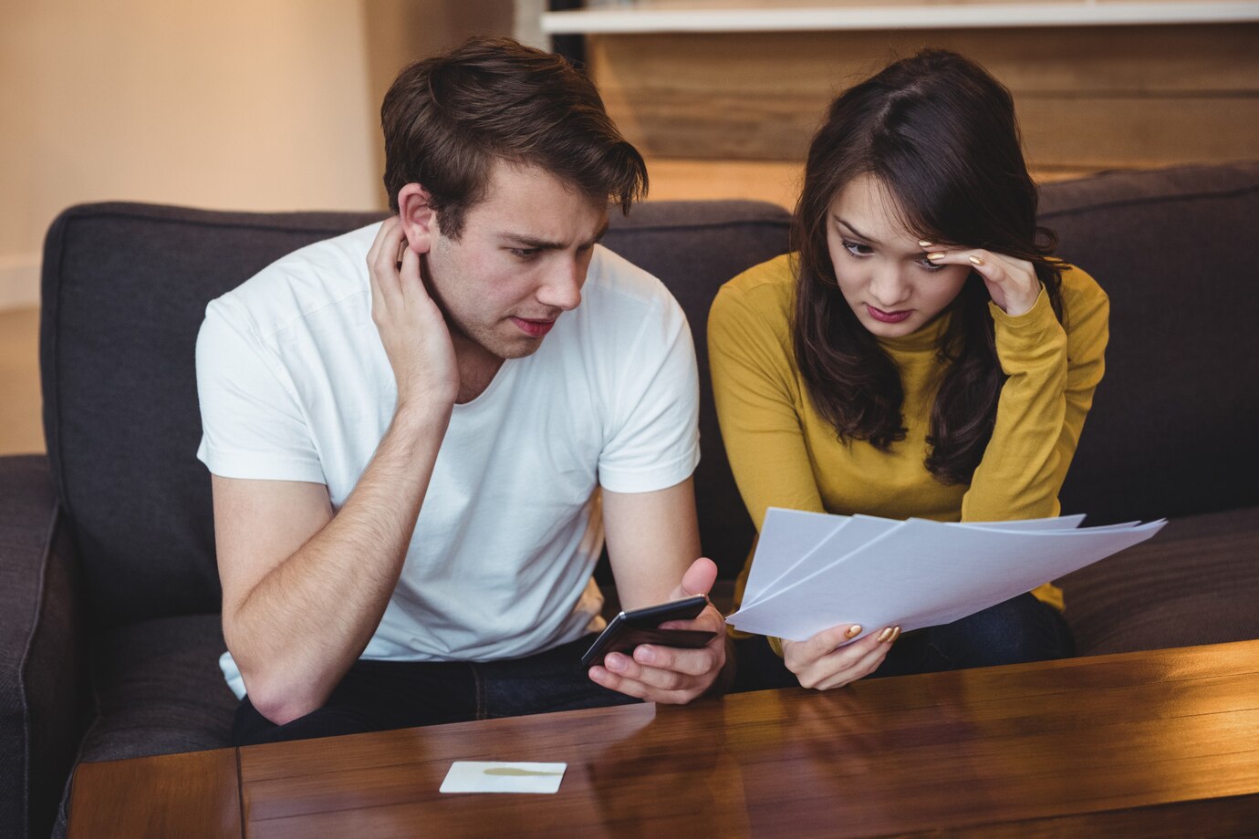A couple sits on a couch, examining papers and a phone, showing expressions of concern and concentration in a cozy setting.