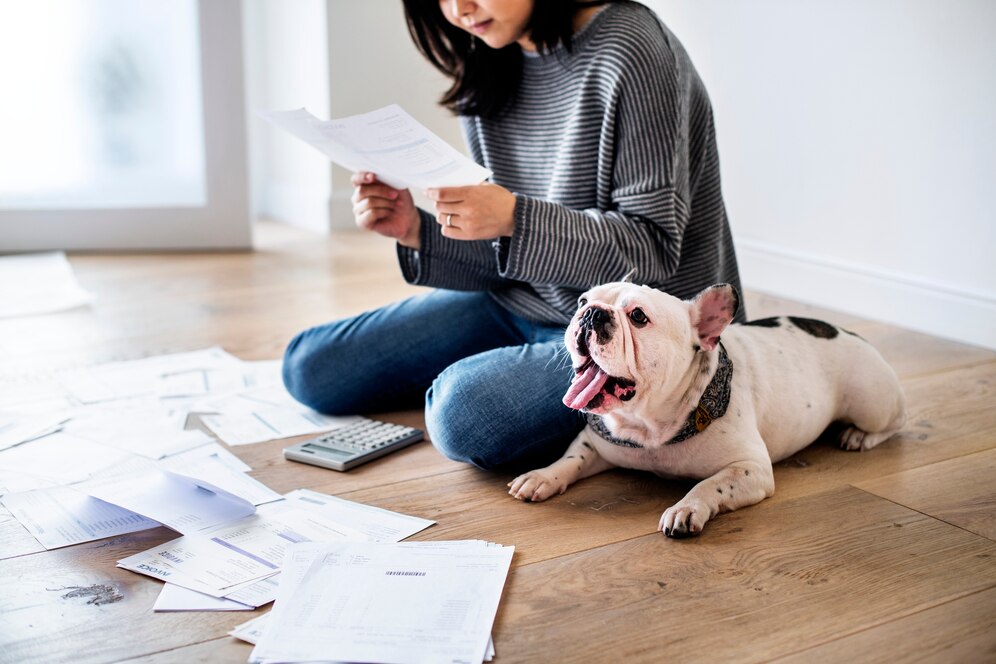 A woman sits on the floor surrounded by papers, reviewing documents while her yawning dog relaxes beside her.