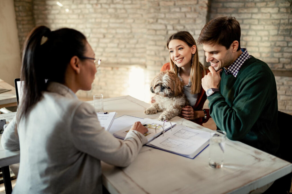A group discussion at a table, featuring a woman in a suit and a couple with a small dog, surrounded by papers and a laptop.