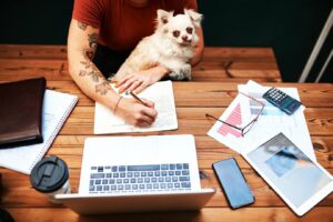 A person writes in a notebook while sitting at a wooden table, surrounded by a laptop, papers, a phone, and a coffee cup. A small dog sits nearby.