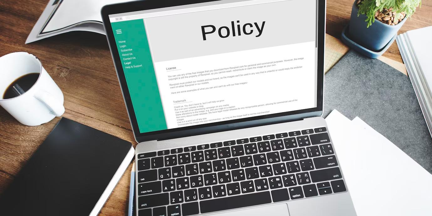 A laptop displaying a Policy page with a coffee mug and notebook on a wooden desk, surrounded by paperwork and a plant.
