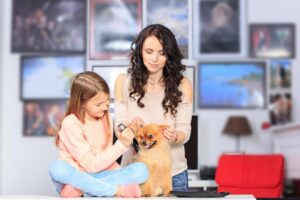 A woman and a girl groom a fluffy Pomeranian dog in a cozy room adorned with various artwork and a red couch.