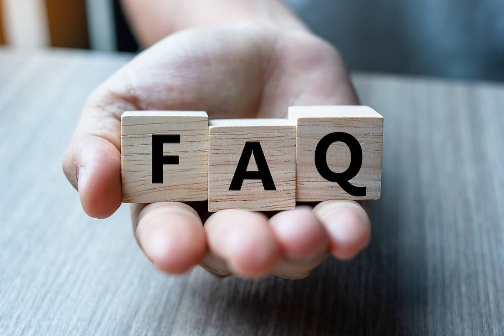 A hand holds wooden blocks spelling FAQ, representing frequently asked questions, against a light wooden background.