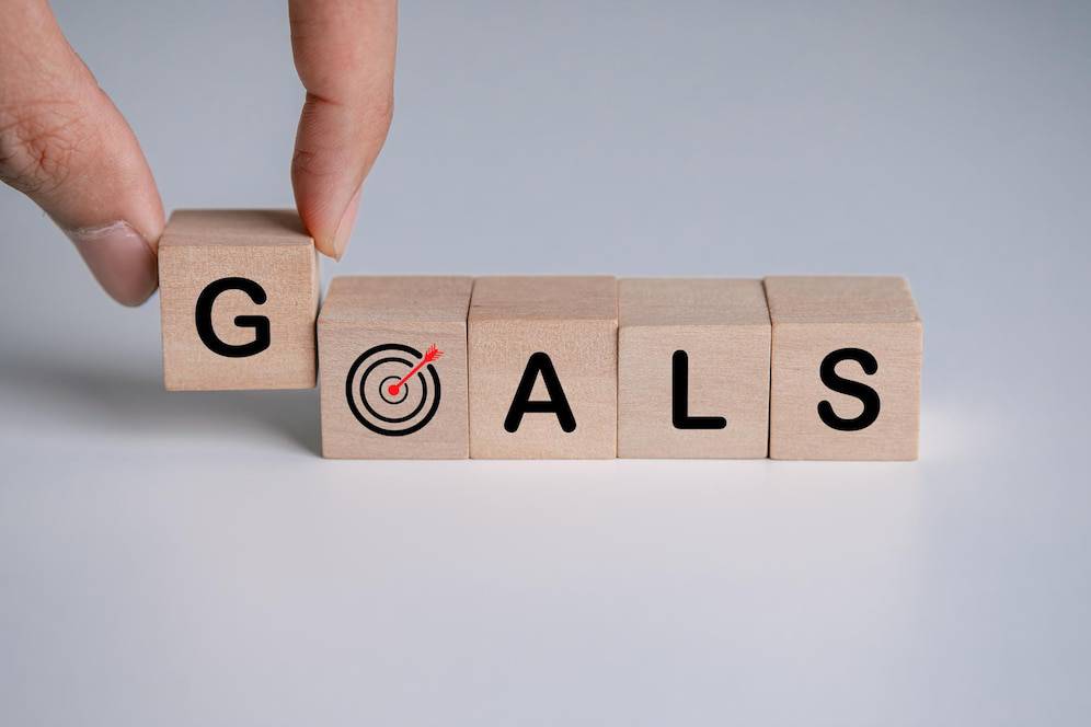 A hand places a block with a target icon beside wooden blocks spelling GOALS on a light background, symbolizing goal setting.