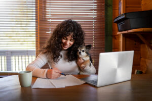 A person writes notes while holding a small dog, with a laptop and a cup of coffee on a wooden desk, sunlight streaming through blinds.