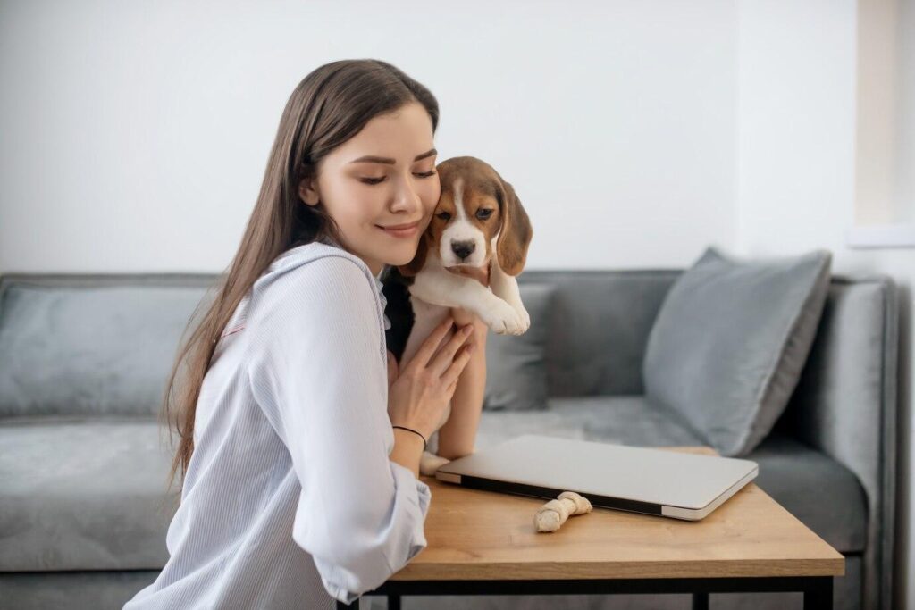 A woman gently holds a small dog close, seated at a table with a laptop, in a cozy living room setting.