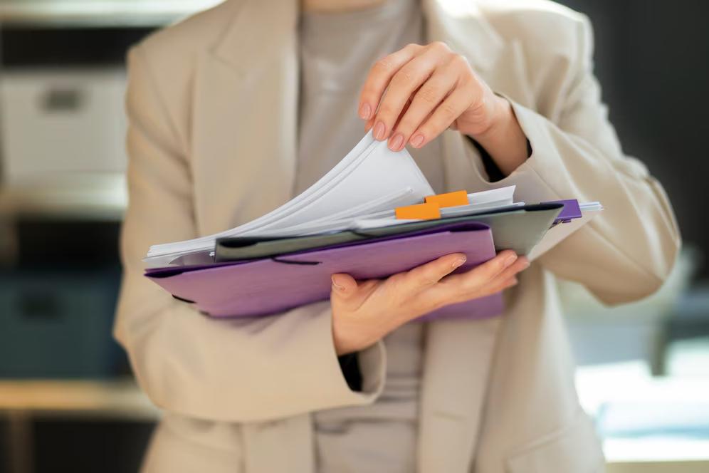 A person in a beige blazer holds a stack of documents and folders, flipping through the papers in a professional setting.