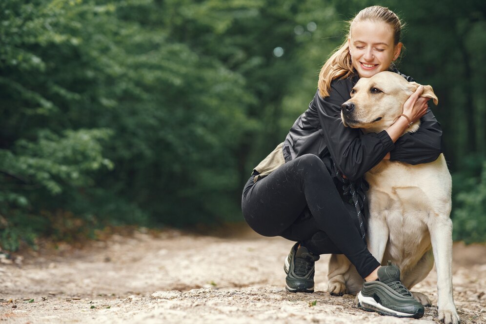 A woman hugs her yellow Labrador retriever in a lush green forest, expressing love and companionship on a dirt path.
