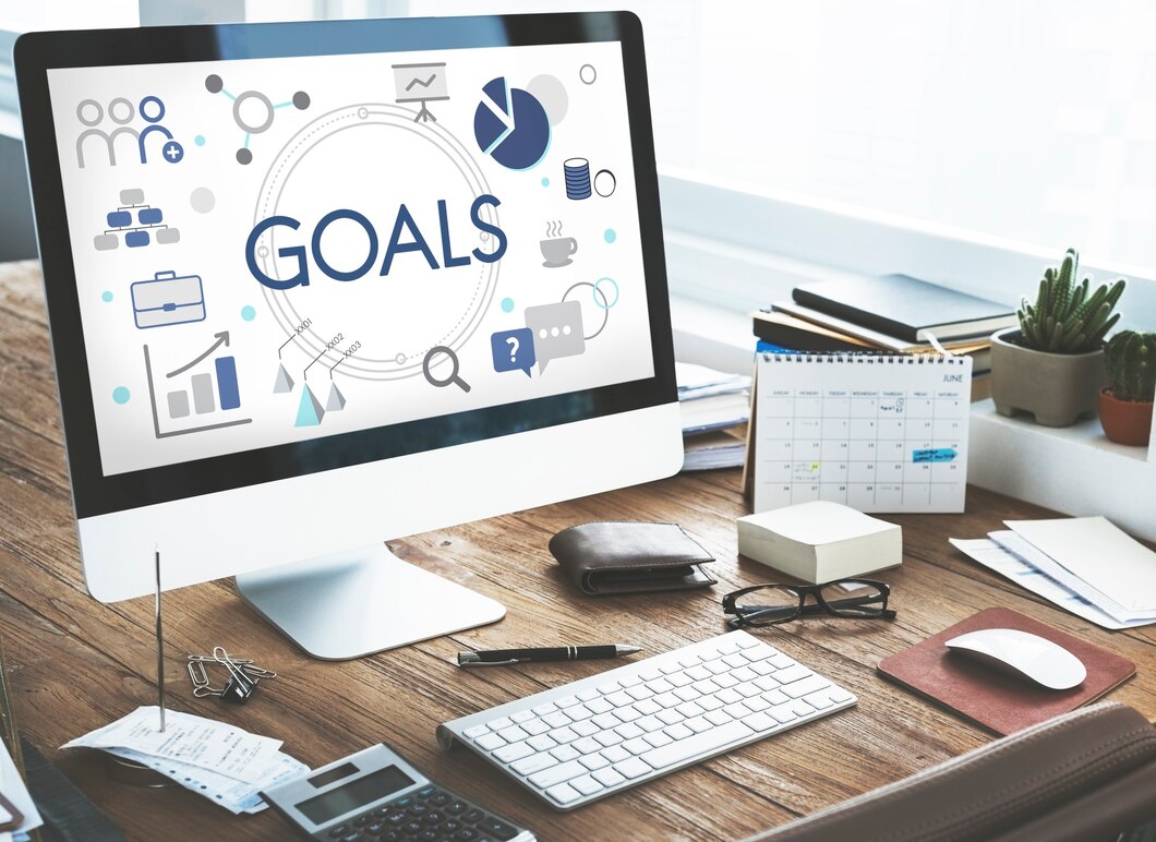 A computer monitor displays the word GOALS surrounded by icons representing business and planning, on a wooden desk with office supplies.
