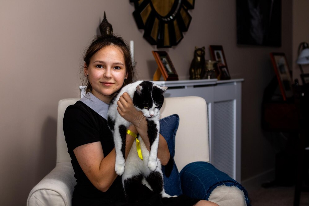 A person sitting on a couch holds a fluffy black and white cat, surrounded by a cozy living room with decorative elements.
