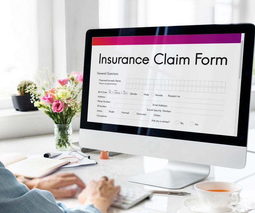 A computer screen displays an insurance claim form, while a person types on a keyboard with a cup and flowers on the desk. 