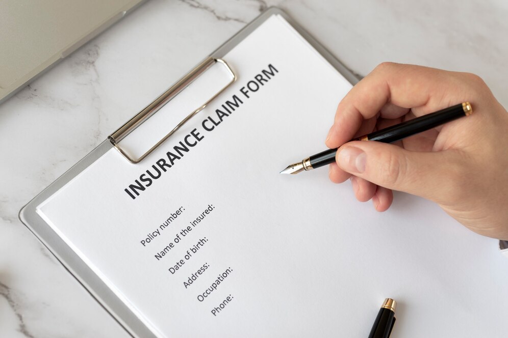 Hand holding a fountain pen above an insurance claim form on a clipboard, with sections for personal details visible.