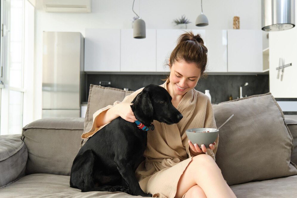 A person in a robe sits on a couch, holding a bowl and sharing a cozy moment with a black dog. Bright, modern kitchen in background.