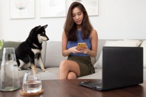 A woman sits on a couch, holding a yellow phone, while a black and tan dog watches her. A laptop and water glass are on the table.