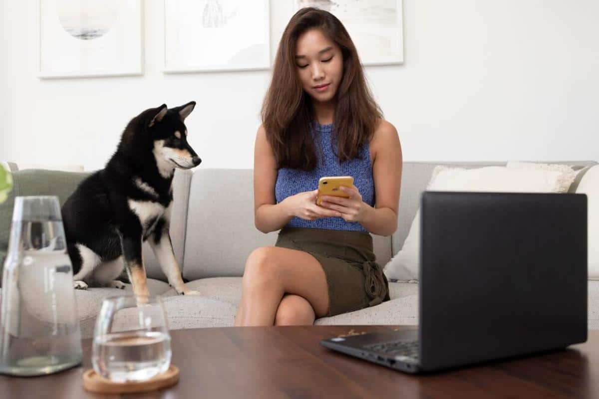 A woman sits on a couch, holding a yellow phone, while a black and tan dog watches her. A laptop and water glass are on the table.