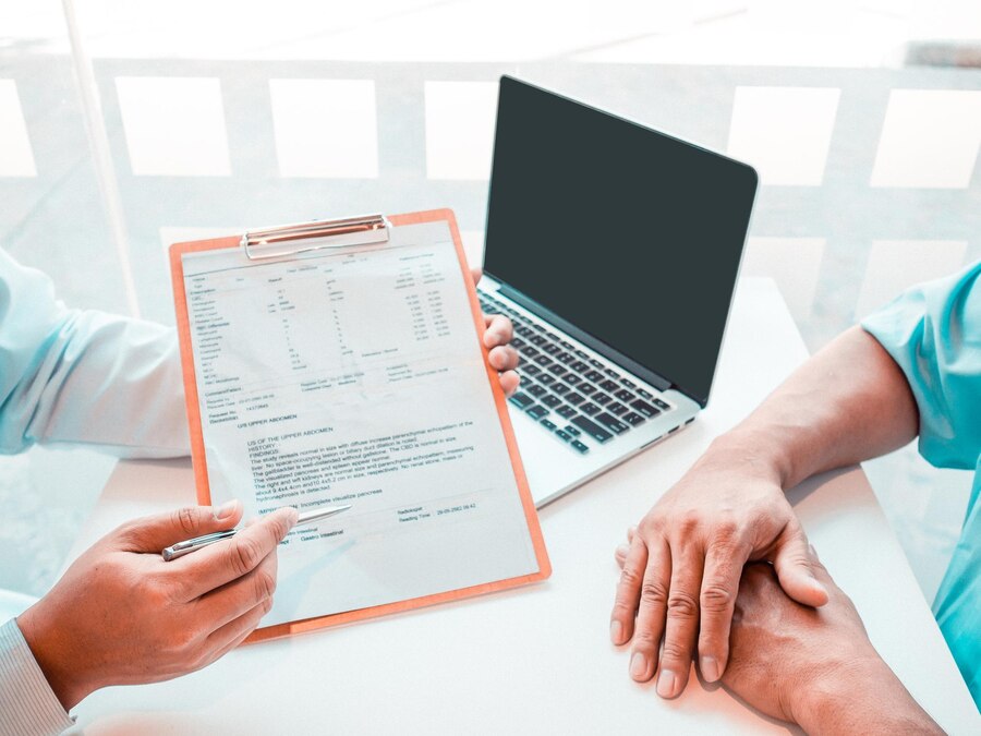 A person holds a clipboard with a document while another person rests their hands on the table next to a closed laptop.