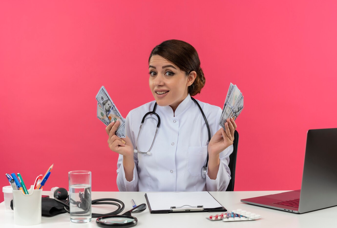 A person in a white lab coat holds bundles of cash, sitting at a desk with a stethoscope, water glass, and a laptop against a pink background.