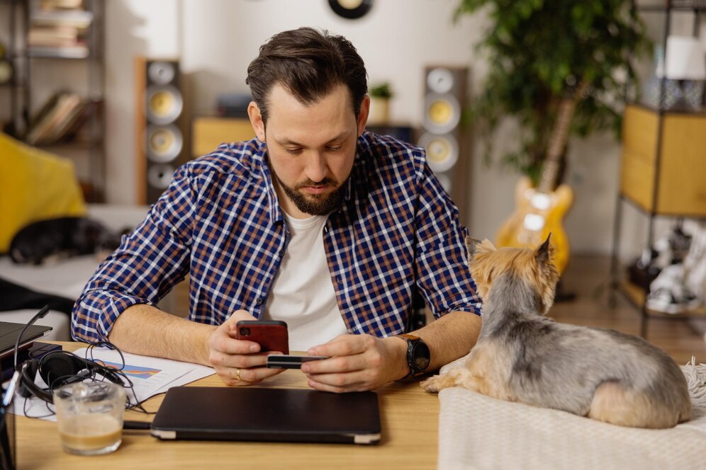 A person in a checked shirt sits at a desk, using a phone, while a small dog rests beside them, surrounded by various devices and paperwork.