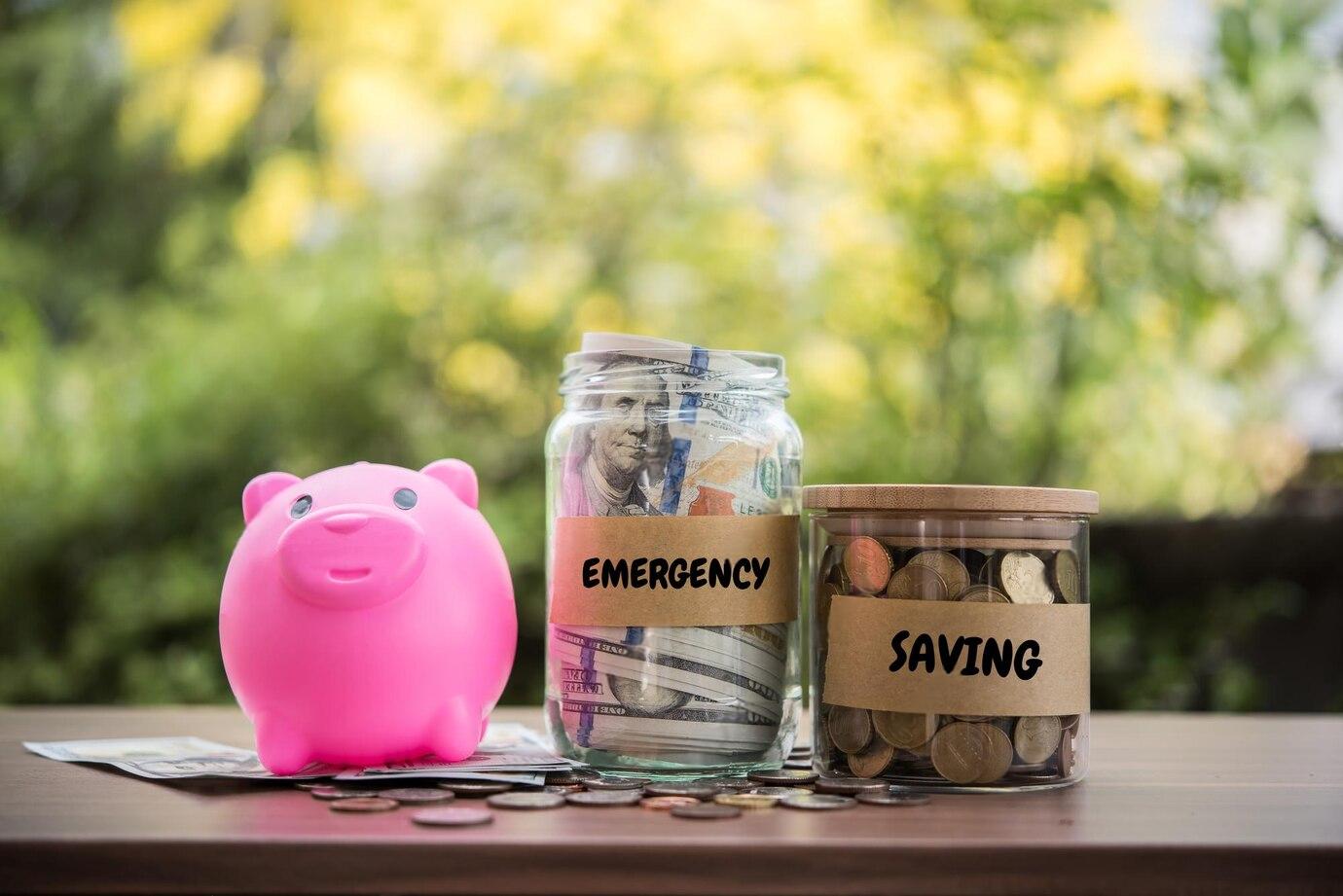 A pink piggy bank sits next to two labeled jars, one for Emergency cash and the other for Saving coins, on a wooden table.